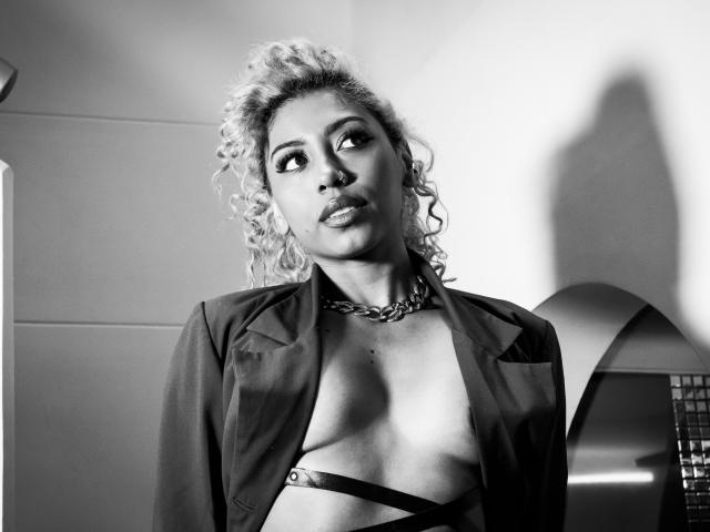 Black-and-white photo of LilithWalker with curly blonde hair, nose ring, chain necklace, open jacket, strappy top, looking up, shadow on wall, visible breasts, confident expression.