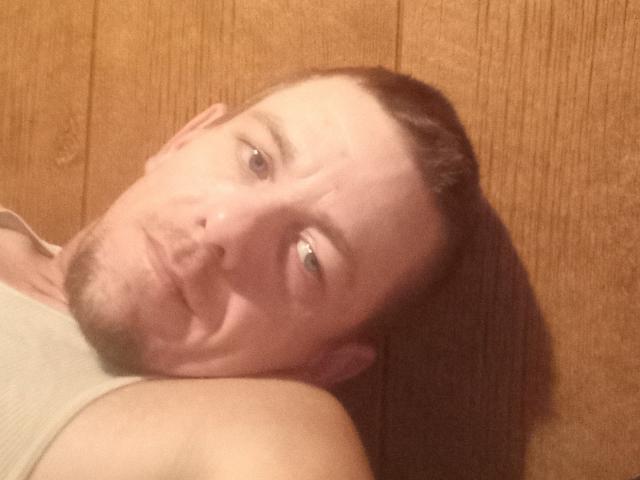 CountryFun has a pale face with a thin beard, blue eyes, and brown hair in a ponytail, lying down with his head tilted, wearing a beige tank top.
