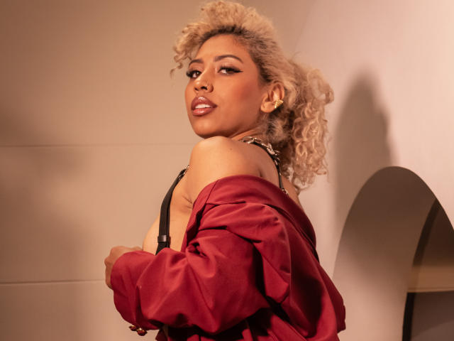 LilithWalker with curly blonde hair, wearing a red shirt off-shoulder, black strap dress, looking over shoulder, showing her upper chest and smiling, with subtle makeup and gold earrings.