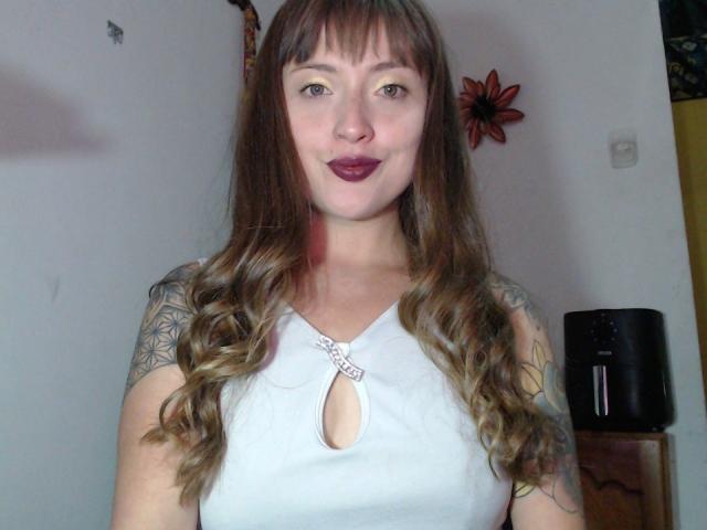 Guren69 with long brown wavy hair, tattoos on arms, wearing a white sleeveless top with a keyhole cutout, and dark purple lipstick. Boobs visible through keyhole.