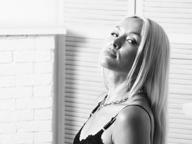 Black-and-white photo of AvaGreenx with long blonde hair, looking up with a slight smirk, wearing a lace bra, chain necklace, and showing her small tits.