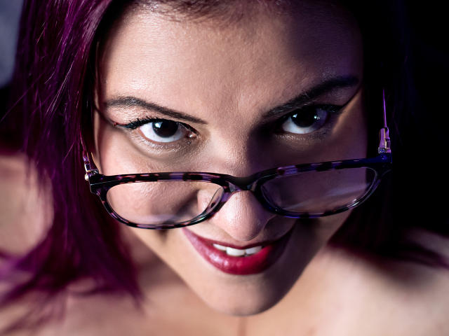 Sammy Owens mit lila Haaren, Schildpattbrille und rotem Lippenstift, lächelt in die Kamera, ihre Brüste sind leicht sichtbar, sie wirkt sexy und selbstbewusst..