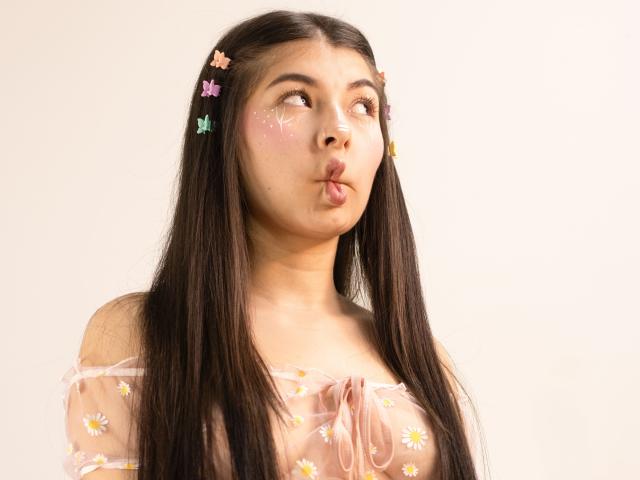 Abbie69 with long dark hair, wearing a sheer pink top with daisy patterns, puffy pink cheeks, and flower hairpins, puckering her lips. Her tits are partially visible.