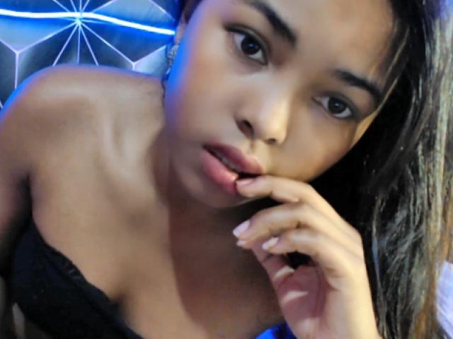 MirahX with dark brown skin, long black hair, and slightly parted lips, touching her bottom lip with her finger, wearing a black lace bra, looking into the camera.