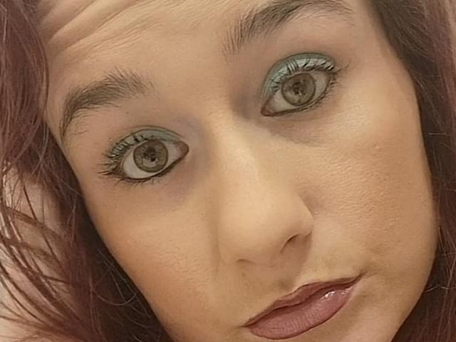 GoddessKatty69's face, showing brown eyes with greenish eyeshadow, brown eyebrows, and red lipstick, with her head slightly tilted and her lips parted in a subtle, sexy pout.