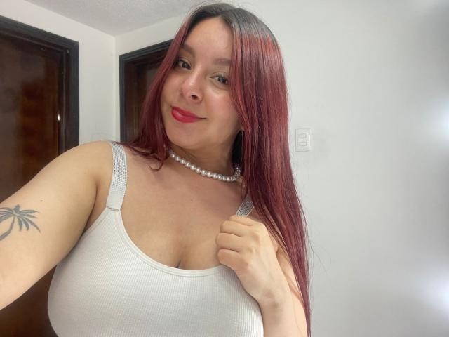 EmaJames has red hair, wears a white tank top, pearl necklace, and smiles slightly with red lipstick. Her right hand holds the tank top, showing a palm tree tattoo.