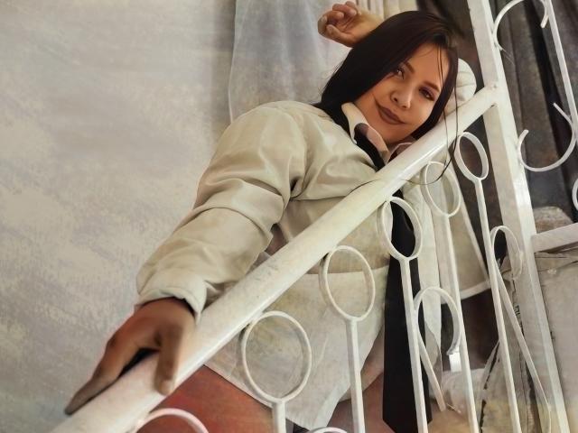 AvaChanne with brown hair, wearing a white shirt and black tie, leans against a white metal railing, her hand gripping it, her face slightly tilted downward, looking relaxed.