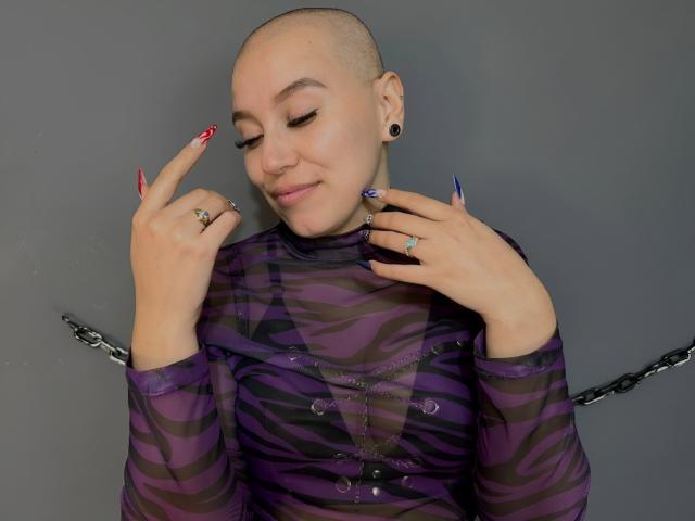 HotAndrea69 mit einem rasierten Kopf, trägt ein durchsichtiges, lila Top mit Zebra-Muster, zeigt bunte, lange Nägel, schwarze Ohrstecker und Ringe, mit einem kleinen Lächeln und geschlossenen Augen..