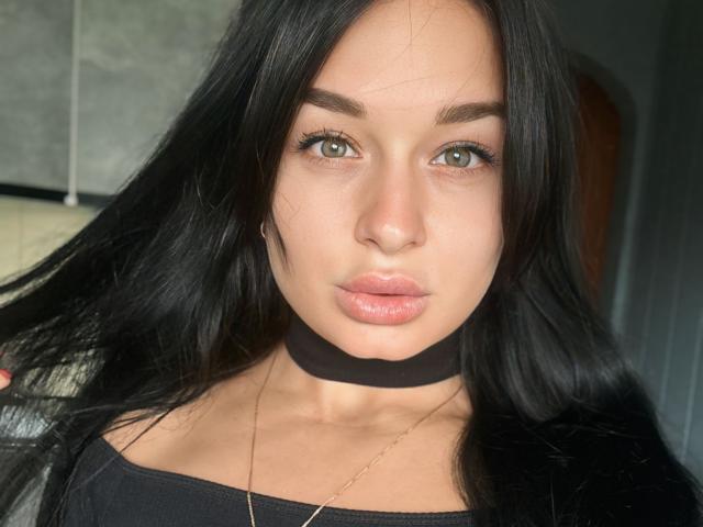 MaryLoveX with long black hair, green eyes, and pink lips, wearing a black choker and black top, has her hair pulled to the side, showing a serious expression.