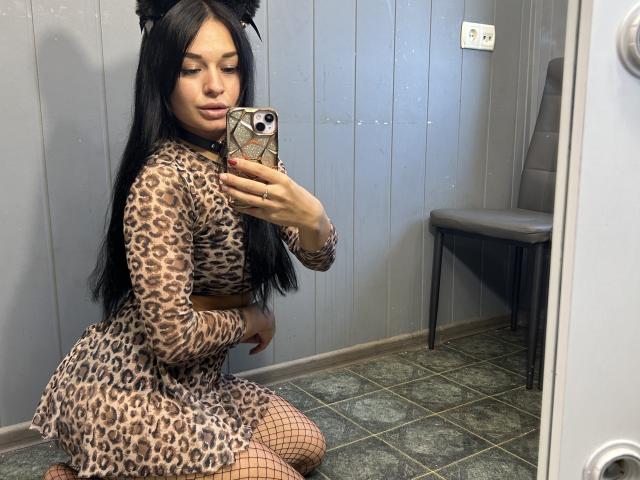 MaryLoveX kneeling, wearing leopard print dress, fishnet stockings, black cat ears, long black hair, holding phone, mirror selfie, green tiled floor, grey wall, grey chair in background.