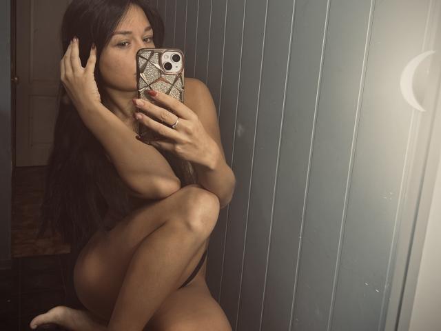 MaryLoveX with long dark hair, naked, squatting, holding a phone with a snake-pattern case, covering her tits with her arm, taking a mirror selfie, showing her ass.
