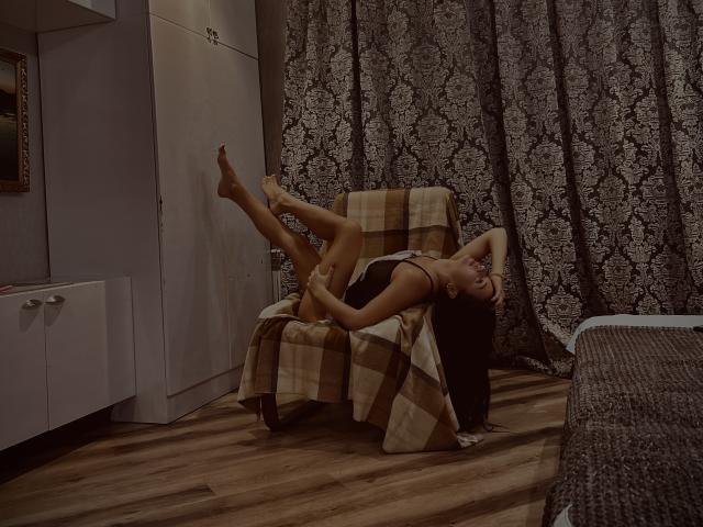 MaryLoveX is lying on a chair, her legs in the air, wearing a black thong, with her ass and tits visible, in a dimly lit room with wood floors.