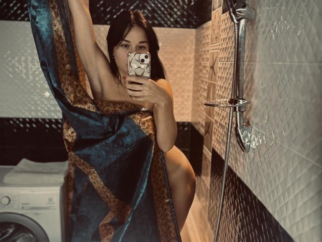 MaryLoveX with dark hair, holding phone, wearing a blue and gold patterned towel, showing her bare ass, in a tiled shower, taking a selfie.