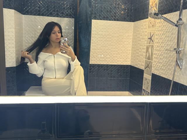 MaryLoveX with long black hair, wearing a white off-shoulder dress and holding her hair, sitting in a tiled bathroom, taking a mirror selfie with a smartphone.