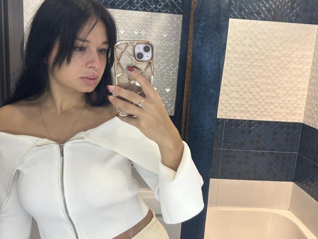 MaryLoveX with long black hair, wearing a white off-shoulder ribbed top, showing her small tits, holding a phone with a geometric case, in a bathroom mirror selfie.