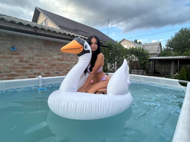 MaryLoveX with long black hair, smiling, sitting on a white inflatable duck in a blue-tinted hot tub, wearing a pink bikini, showing her ass.