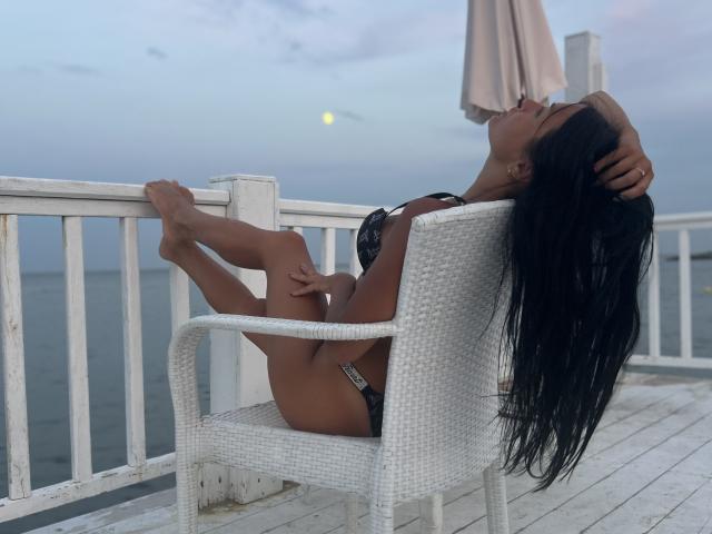 MaryLoveX with long black hair, dark skin, and small black bra and panties, lounging on a white wicker chair, kissing a beige umbrella, feet up, hands on head.