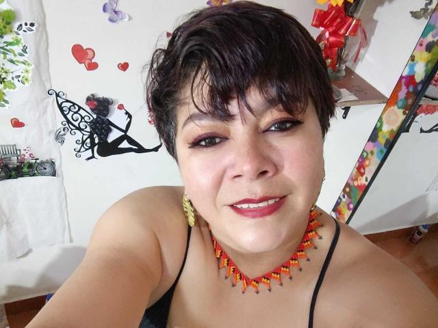 Angellovelly with short dark hair, wearing a black spaghetti strap top and orange-red bead necklace, smiling at the camera, showing a cute face and small tits.