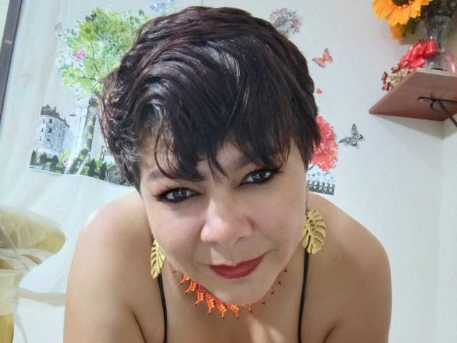 angellovelly with short black hair, wearing a black strap top, orange necklace, and sunflower earrings, smiling with red lipstick, looking down at the camera. Her tits are visible.