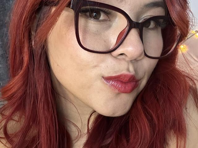 MelisaBrunette with red hair, wearing black glasses, has shiny pink lips and a slight pout, showing subtle makeup and a cute, sexy face. Her ass is not visible.