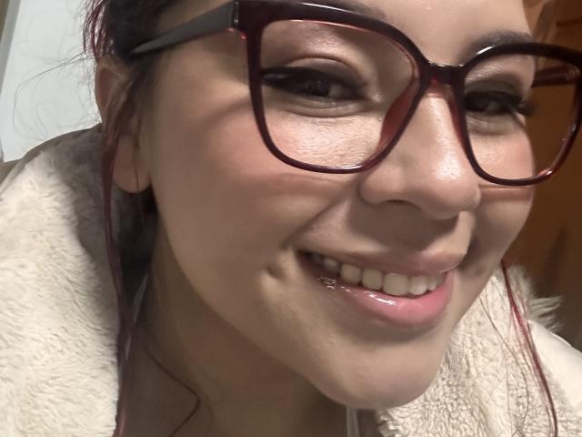 MelisaBrunette with glossy pink lips, shiny teeth, and a cute smile, wearing red glasses and a white fuzzy jacket, has a slight nose crease and auburn hair.