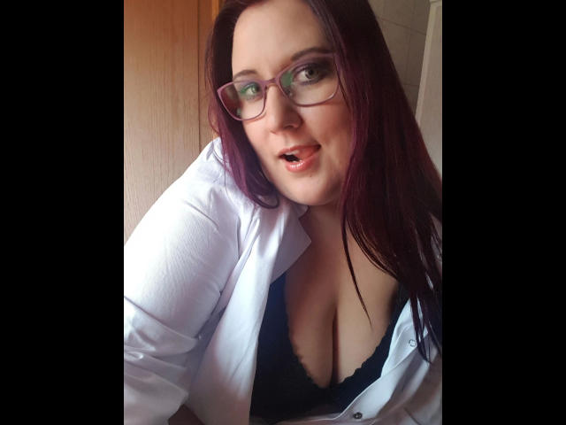 JosySweet, mit Brille, rotem Haar und offenem, weißem Hemd, unter dem ein schwarzer BH und tiefer Ausschnitt zu sehen sind. Sie blickt mit leicht geöffnetem Mund in die Kamera..