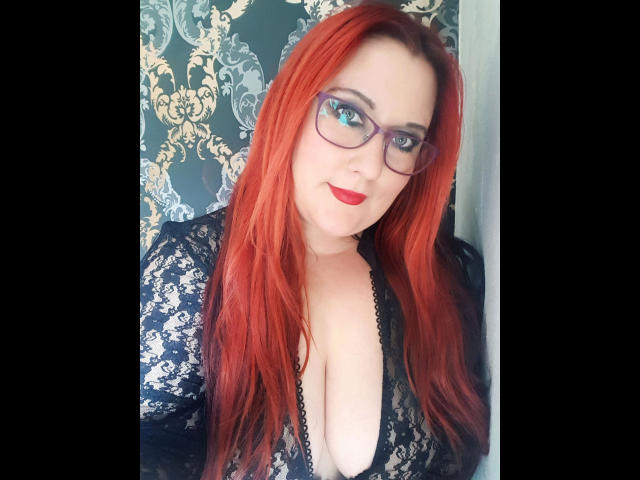 JosySweet : Cheveux roux, lunettes et rouge à lèvres rouge, portant un haut en dentelle avec un décolleté plongeant, montrant ses seins. Elle sourit discrètement..