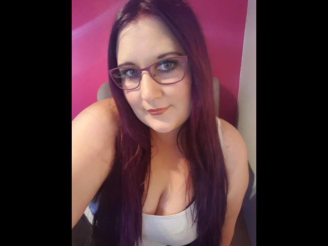 JosySweet – heiß, mit langen roten Haaren, Brille und einem weißen Tanktop, das ihre großen Brüste zeigt, lächelnd in die Kamera. Rosa Hintergrund..