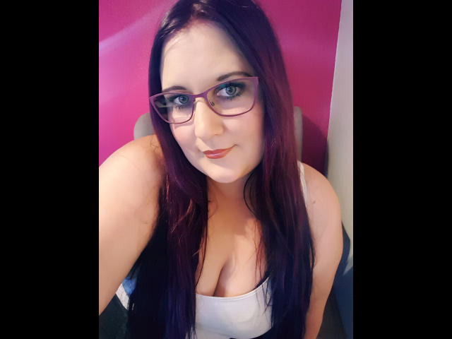 JosySweet-hot with glasses, long dark red hair, and a white tank top showing her big boobs, smiling slightly at the camera. Pink background.