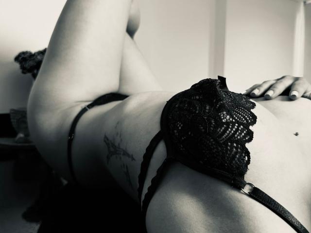 Le soutien-gorge en dentelle noire d'Arenne est porté sur ses petites seins, sa jambe est levée, révélant un tatouage sur sa hanche, et sa main repose sur ses fesses. Photo en noir et blanc..