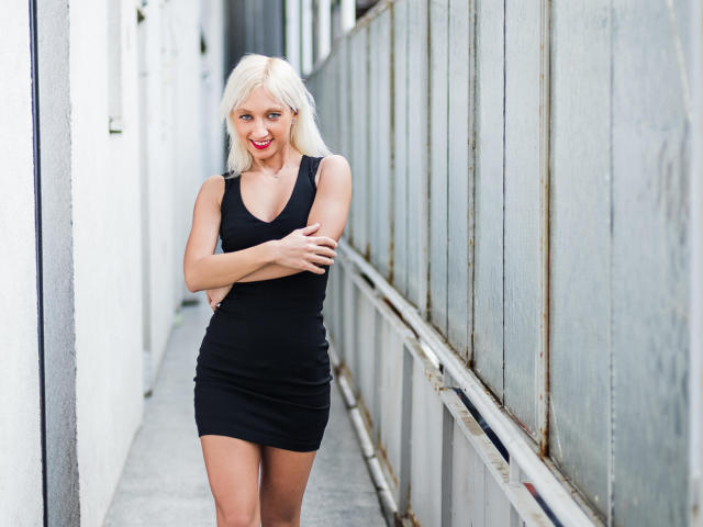 NatalieXBitton, con i capelli biondi, un vestito nero e rossetto rosso, è in piedi in un corridoio stretto, con le braccia incrociate, mostrando i suoi piccoli seni e glutei, sorridendo con un viso carino..
