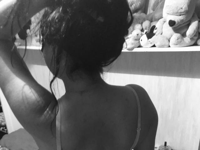 Black-and-white photo of QueridaCalina with her back to the camera, hands in her messy dark hair, wearing a thin-strapped bra, showcasing her ass and a slight tattoo on her arm.