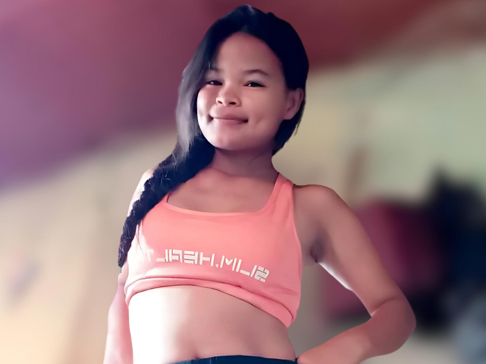 PatrikStar69, smiling, wearing a pink sports bra, exposing a flat belly and small tits, with long black hair in a braid, standing confidently with hands on hips.