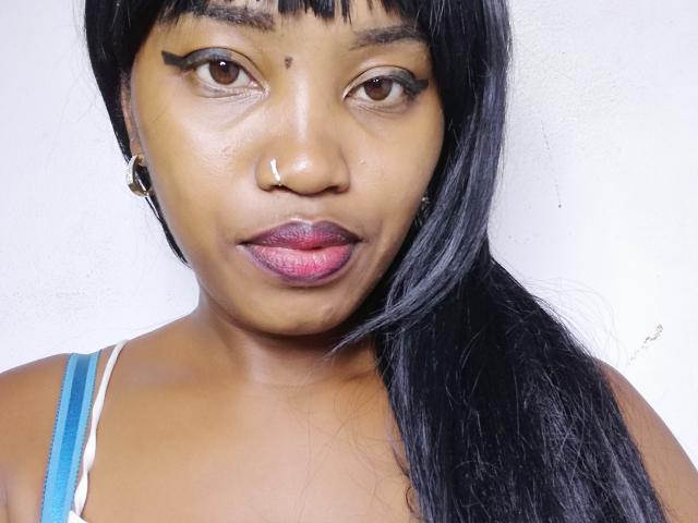 ChaimaBella's face, dark brown skin, black eyeliner, nose ring, straight black hair, pink lipstick, blue and white strap, slight makeup, subtle smile, white wall background.