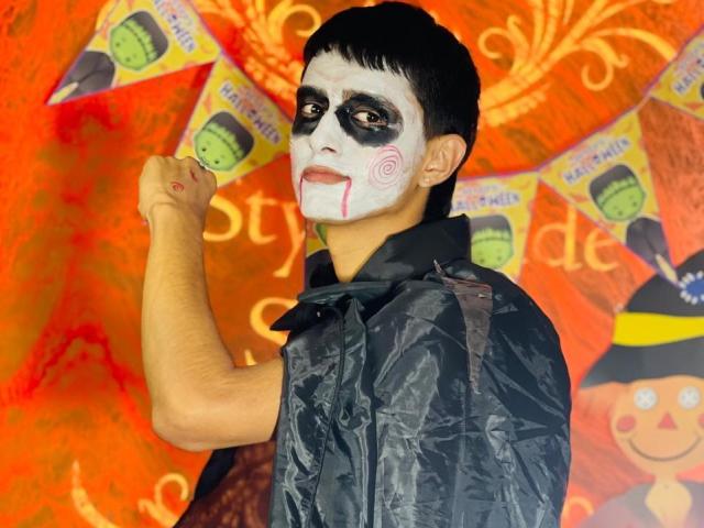 NathanMaxime con il viso dipinto di bianco, trucco nero intorno agli occhi, labbra rosse, un panno nero, un pugno alzato, capelli neri corti, sfondo arancione e rosso, una maschera verde e irregolare sullo sfondo..