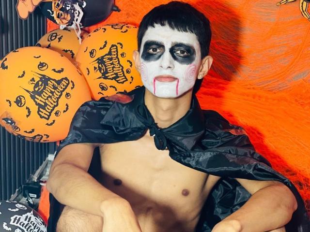 NathanMaxime, torse nu, avec du maquillage noir sur le visage, du blanc autour des yeux et de la bouche, une cape noire, des cheveux courts et foncés, des ballons orange et noirs en arrière-plan..