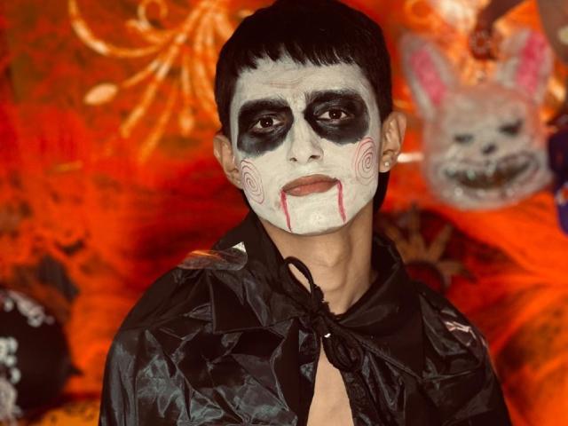 NathanMaxime, mit weißer Gesichtsfarbe, schwarzem Augen-Make-up, rotem Lippenstift und einer blutenden Nase, trägt einen glänzenden schwarzen Plastikmantel, hat kurzes schwarzes Haar und einen ernsten Gesichtsausdruck..