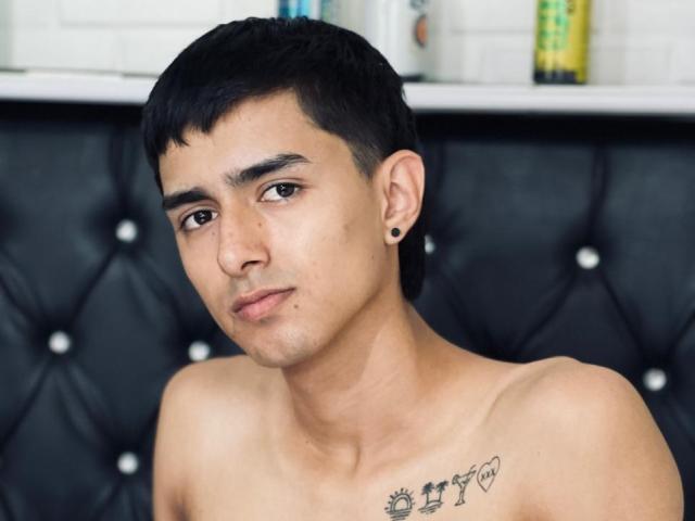 NathanMaxime, senza camicia, con capelli neri corti, piccoli orecchini neri e un tatuaggio sul petto destro, ha un'espressione neutra. Lo sfondo è in pelle nera con una texture simile a quella di un tappeto..