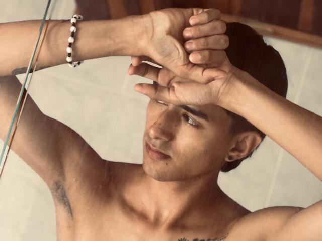 NathanMaxime, senza camicia, con le braccia incrociate sulla testa, ha un braccialetto al polso destro, capelli scuri e un leggero sorriso. I peli sotto le ascelle sono visibili. Ha i piercing alle orecchie..
