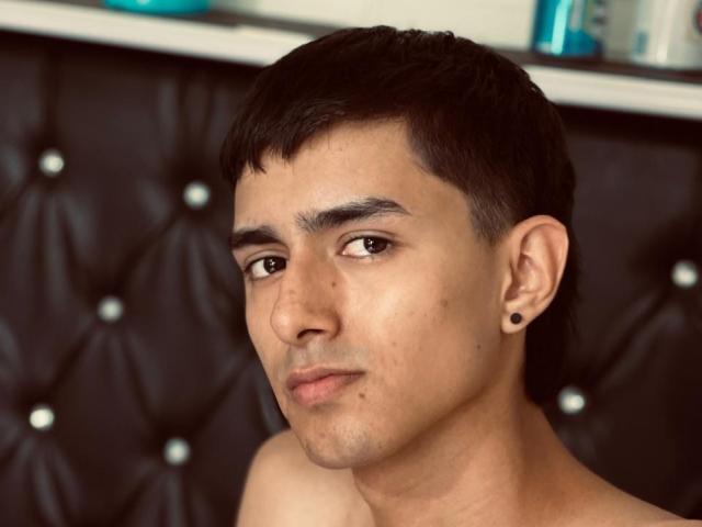 NathanMaxime tiene el pelo corto y negro, ojos marrones y un pequeño piercing negro en la oreja derecha. Está sin camisa y tiene una expresión neutra..