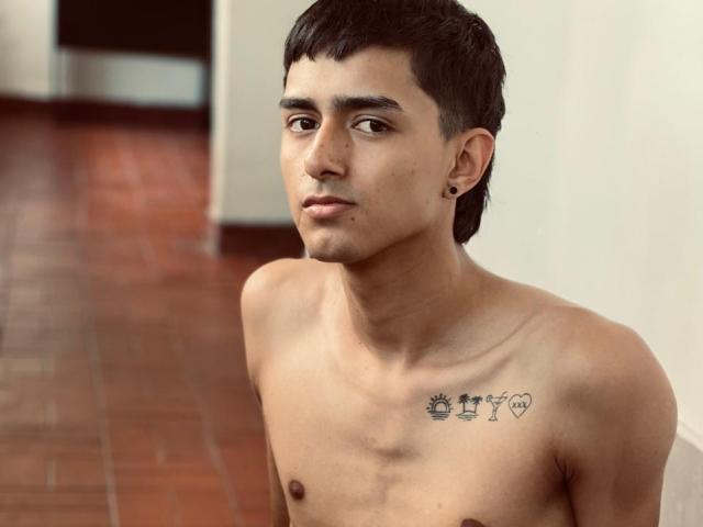 NathanMaxime sem camisa, com cabelo preto curto, pequenas tatuagens no peito e piercings nas orelhas, olhando para baixo com uma expressão neutra. Olhos castanhos, pele marrom clara, chão de azulejos marrons..