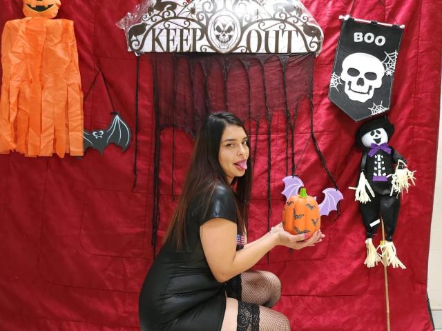 SaraAndMarilyn69 vestindo um vestido de couro preto, meias de rede, segurando uma abóbora, língua de fora, ao lado de decorações de Halloween, um vestido laranja, um morcego, um crânio e um fantasma em um tecido vermelho..
