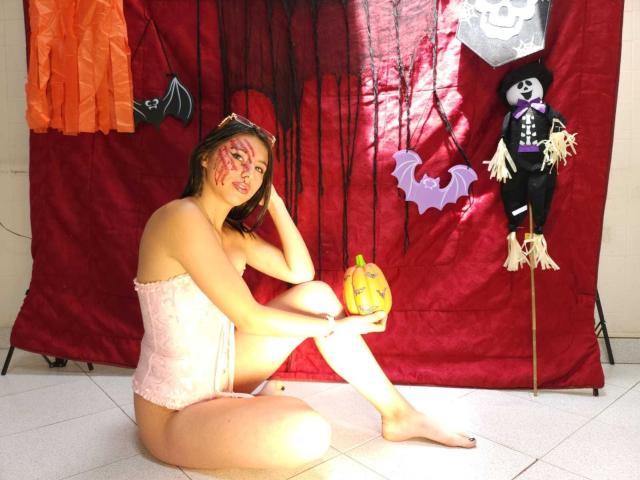 KloeDekerr sits on floor, wearing pink lace corset, no pants, holding pumpkin, face painted, orange and purple bat decorations, and creepy doll on stick, in sunlit room.