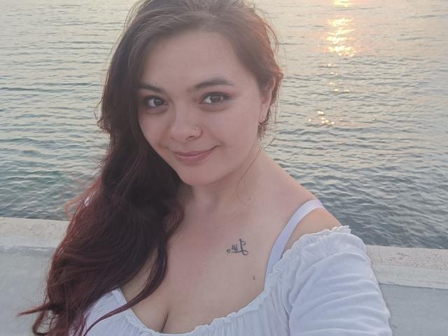 KatherinLatin with long dark red hair, white off-shoulder top, showing her tits, small tattoo above her right breast, smiling, standing by water at sunset.
