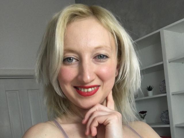 NatalieXBitton ha i capelli biondi, gli occhi azzurri, il rossetto rosso e un sorriso carino, con un dito appoggiato sul mento. Indossa una maglietta rosa e ha un aspetto sexy..