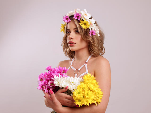SkyCyrus with wavy blonde hair, wearing a flower crown, holding colorful flowers (pink, white, yellow), in a white halter top, looking slightly down, with a neutral expression.