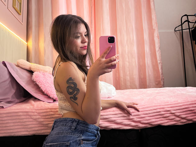 SunBlonde with a snake tattoo on her arm, taking a selfie in a pink room, wearing a lace bra and blue jeans, holding a pink phone, showing her tits.
