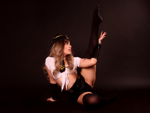 SkyCyrus with long blonde hair, wearing a pilot hat, tied shirt, black shorts, and thigh-high socks, raising one leg, showing her tits and tattoo on her arm.