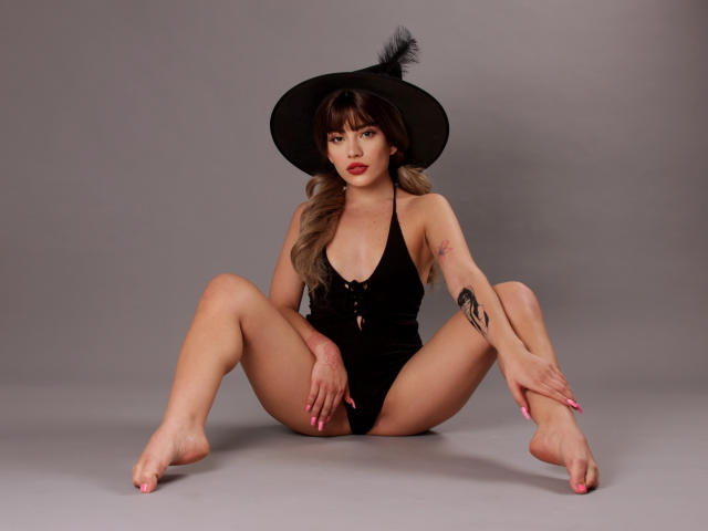 SkyCyrus, sitting with legs spread, wearing a black hat with a feather, black one-piece, showing her tits, tattoo on right arm, pink nails, bare feet, grey background.