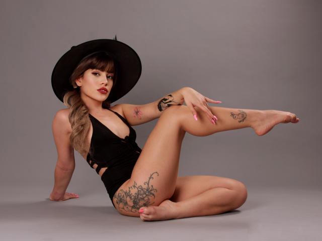 SkyCyrus sitting on the floor, legs up, wearing a black hat and revealing black one-piece, showing tattoos on arms and thigh, with her boobs and pussy subtly visible.